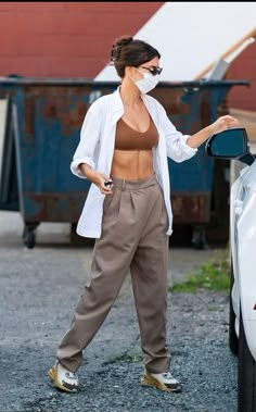 a woman in brown pants standing next to a white car and wearing a face mask