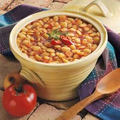 a yellow bowl filled with beans next to a wooden spoon and tomato on a table