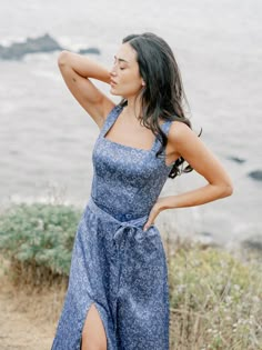 a woman in a blue dress standing by the ocean with her hand on her head