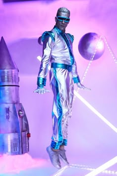 a man dressed in silver and blue standing next to a rocket ship