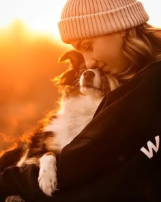 a woman holding a dog in her arms with the sun setting behind her and she is wearing a beanie