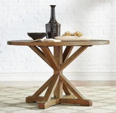 a wooden table with a vase and bowl on top of it in front of a white brick wall