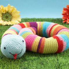 a crocheted snake laying on the grass with flowers in the backgroud