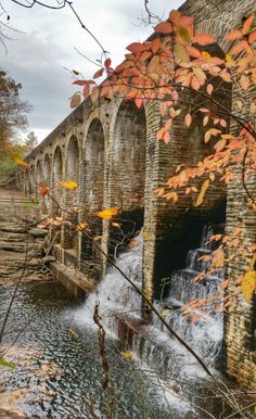 an old stone bridge over a river with fall leaves on the trees and water running under it