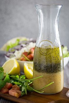 a glass pitcher filled with dressing next to a bowl of salad and lemon wedges