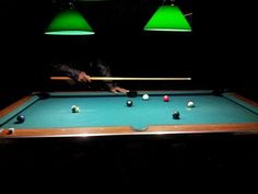 a pool table with two green lamps hanging from it's sides and one person holding a cue