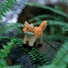 a toy fox standing on top of a rock surrounded by ferns and other greenery