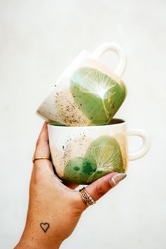 two coffee mugs with leaves painted on them are being held by a woman's hand