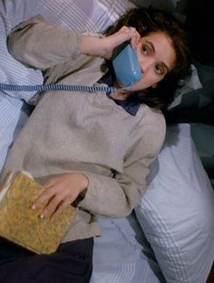 a woman laying in bed with a blue mug on her head while talking on the phone