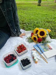 a person sitting at a table with some fruit and flowers in front of them on the grass