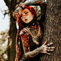 a woman with red hair and tattoos on her body leaning against a tree trunk in the woods