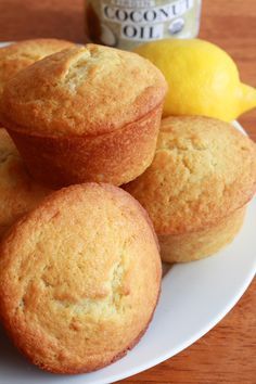 three lemon muffins on a white plate next to two lemons and a jar of condiment