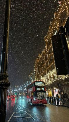 a red double decker bus driving down a street next to tall buildings covered in snow