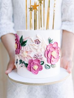 a woman holding a white cake with pink flowers and gold forks on top of it