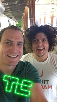 two men sitting at a table with food in front of them and one man holding up a neon sign