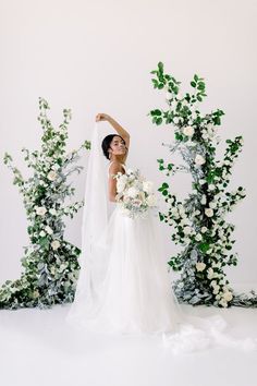 a woman in a wedding dress standing next to an arch with flowers and greenery