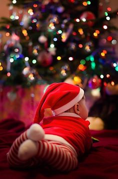 a small baby in a santa hat laying on a bed next to a christmas tree