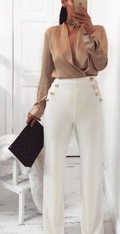 a woman taking a selfie while wearing white pants and a brown top with gold buttons