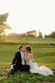 a bride and groom sitting on the grass in front of an open field at sunset