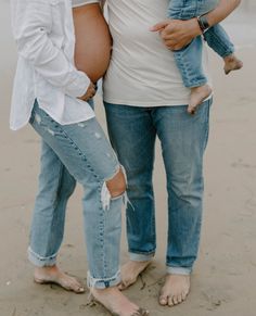 two pregnant women standing next to each other on the beach with their hands in their pockets