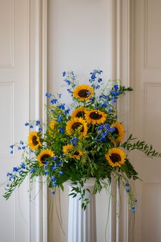 a white vase filled with blue and yellow flowers on top of a wooden floor next to a wall