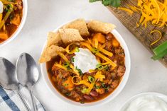 two bowls of chili with cheese, sour cream and tortilla chips on the side