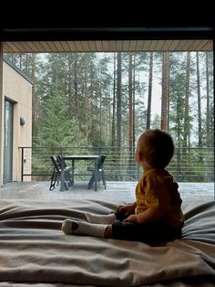 a small child sitting on top of a bed in front of a large window with trees outside