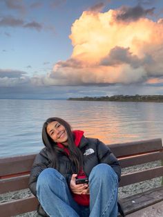 a woman sitting on top of a wooden bench next to the ocean under a cloudy sky