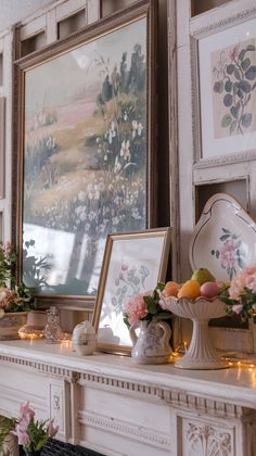 a mantle with flowers, fruit and candles in front of pictures on the wall above it