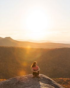 a woman sitting on top of a rock with the sun setting in the distance behind her