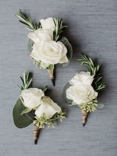 three boutonnieres with white flowers and greenery are arranged on a gray surface