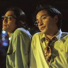 two men wearing glasses and ties standing next to each other in front of a dark background