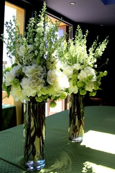 two vases filled with white flowers on top of a green tablecloth covered table