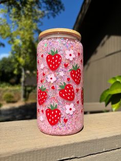 a pink jar with strawberries painted on it sitting on a ledge next to a tree