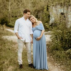 a pregnant couple standing next to each other on a dirt path in the woods with trees behind them