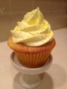 a cupcake with white frosting and yellow sprinkles sits on a cake stand