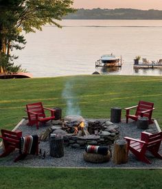 a fire pit surrounded by red chairs in the middle of a grassy area next to a lake