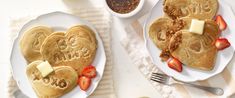 two heart shaped pancakes with butter and strawberries on plates next to a cup of coffee