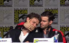 two men sitting next to each other in front of a microphone at a comic convention