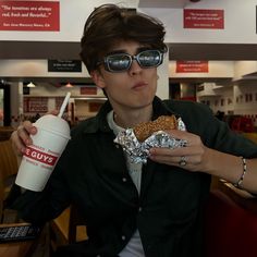 a young man wearing sunglasses and holding a hot dog in his right hand while sitting at a table