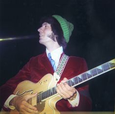 a man in a red jacket and green hat holding a guitar while wearing a beanie