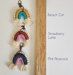 three tasseled key chains hanging from a wooden hook on a white wall next to a sign that says beach girl, strawberry latte, pink peacock and peach