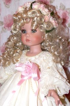 a doll with blonde hair wearing a white dress and pink flower in her hair is sitting on a chair