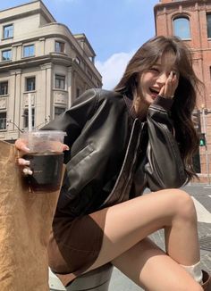 a woman sitting on the ground holding a drink and talking on her cell phone with buildings in the background