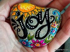 a hand holding a rock with the word joy painted on it and flowers around it