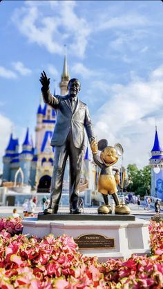 a statue of walt and mickey mouse in front of the castle at disney world with flowers around it