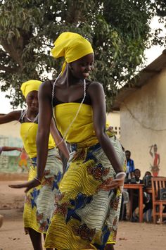 two women in yellow dresses are dancing on the dirt ground while people watch from behind them