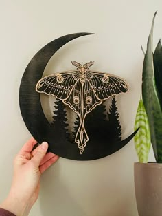 a hand holding a wooden cutout of a moth on a crescent moon with trees in the background