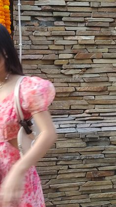 a woman in a pink dress standing next to a stone wall holding a white purse