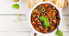 a white bowl filled with food next to some bread and green leafy leaves on the side